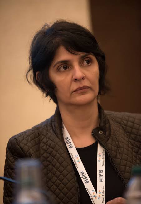 A close-up portrait of Aliya Salahuddin, a freelance writer and documentary filmmaker, wearing a dark quilted jacket and a lanyard for the Google News Initiative with the hashtag #ijf19.
