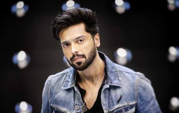 “Fahad Mustafa with styled dark hair and beard wearing light blue denim jacket, looking at camera with confident expression against black background with bokeh lighting effects”
