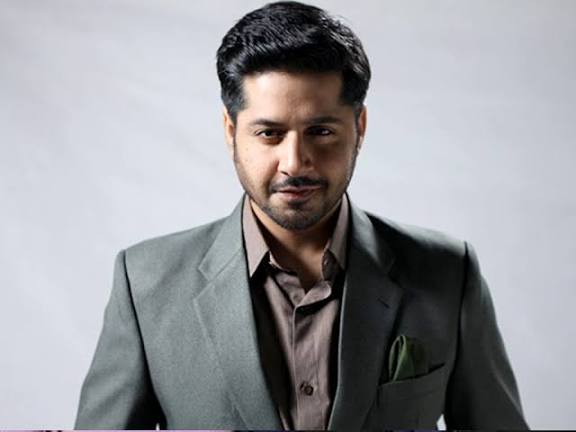 “Imran Ashraf with styled dark hair and beard wearing olive green suit with brown shirt and matching pocket square, looking confidently at camera against white studio background”
