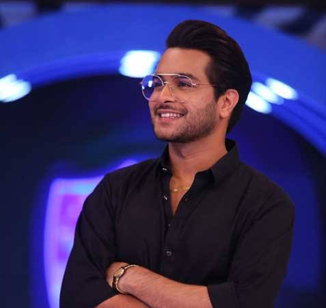 “Man wearing aviator sunglasses and black shirt with arms crossed, smiling on brightly lit television show set with blue lighting”