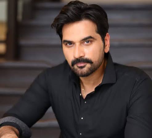 “Man with dark hair and beard wearing black button-down shirt, looking intensely at camera while sitting on outdoor stone steps”