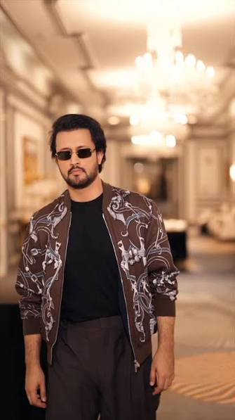 “Man wearing sunglasses and brown patterned bomber jacket over black shirt, standing in elegant hotel corridor with ornate chandelier lighting in background”
