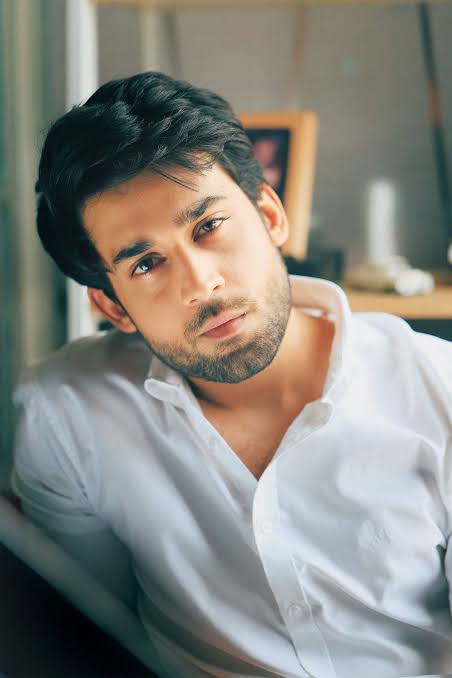 “Man with dark wavy hair and beard wearing white button-down shirt, sitting indoors with hand near neck, looking intensely at camera in soft natural lighting”
