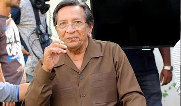 A portrait of veteran Pakistani actor Talat Hussain, who is wearing a brown shirt and glasses, looking at the camera with a thoughtful expression.
