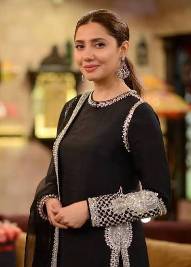 “Woman with hair pulled back wearing elegant black outfit with silver embellished trim and statement earrings, standing in television studio set”