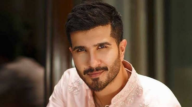  A close-up portrait of Pakistani actor Feroze Khan smiling, wearing a light pink embroidered shirt.
