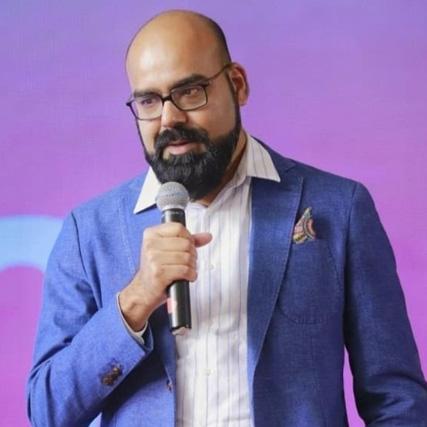 Junaid Akram (Content Creator) with glasses, wearing a blue blazer and a striped shirt, holds a microphone and speaks to an audience.