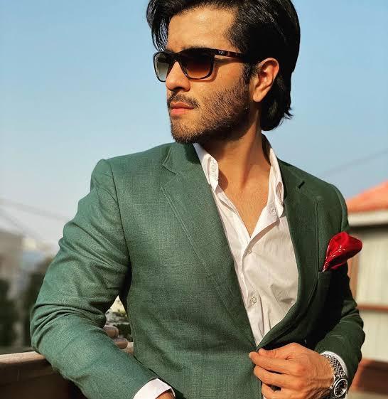 A portrait of Pakistani actor Feroze Khan wearing a dark green blazer, a white shirt, and sunglasses, looking off to the side with a slight smile.