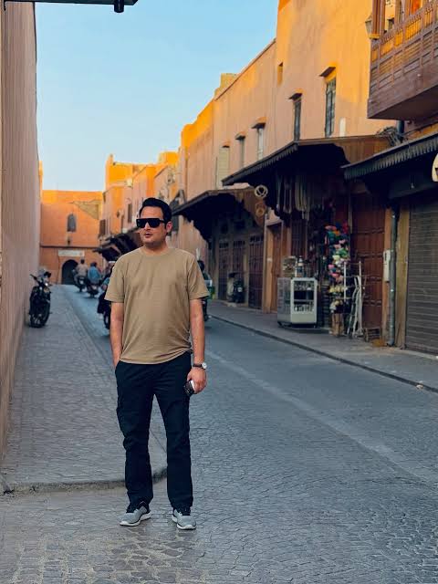 A man wearing a tan t-shirt, black pants, and sunglasses stands on a cobblestone street in a narrow alley in Marrakech, Morocco. The street is lined with buildings and shops with orange-colored walls.
