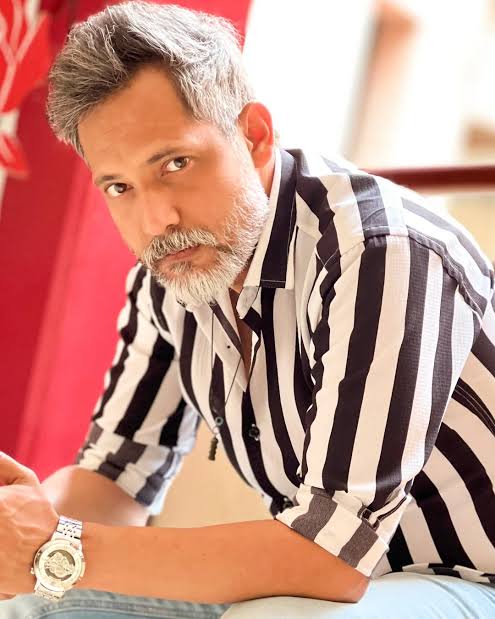  A portrait of Meesam Naqvi, director of the drama “Mayi Ri,” wearing a black and white striped shirt and a watch, with a serious expression.
