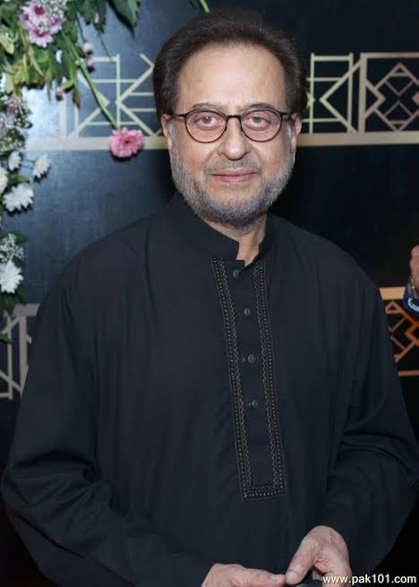 A portrait of Pakistani actor Nadeem Baig wearing a black kurta and glasses, smiling at the camera.

