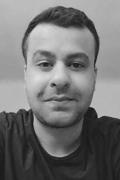 A black and white close-up portrait of a man with short hair and a slight beard, wearing a dark t-shirt.