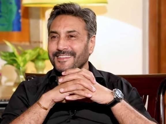 A portrait of Adnan Siddiqui, a Pakistani actor, smiling and looking to the side. He is wearing a black shirt and has his hands clasped.