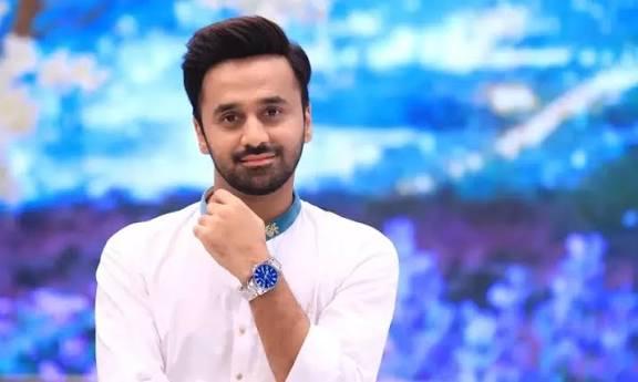 A portrait of Pakistani television host Waseem Badami, wearing a white shirt with a patterned collar, smiling slightly while holding his chin.
