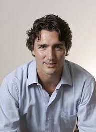 Portrait photograph of Justin Trudeau with dark curly hair, wearing a light blue button-down shirt, leaning forward against a plain background
