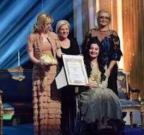 Muniba Mazari receiving an award on stage, seated in a wheelchair, smiling confidently while holding a trophy, dressed in elegant traditional attire.