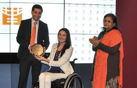 Muniba Mazari receiving an award on stage, seated in a wheelchair, smiling confidently while holding a trophy, dressed in elegant traditional attire.