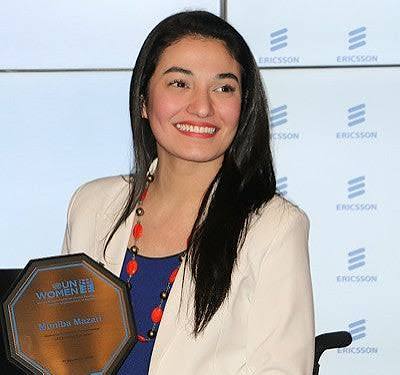 Muniba Mazari receiving an award on stage, seated in a wheelchair, smiling confidently while holding a trophy, dressed in elegant traditional attire.