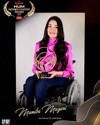 Muniba Mazari receiving an award on stage, seated in a wheelchair, smiling confidently while holding a trophy, dressed in elegant traditional attire.
